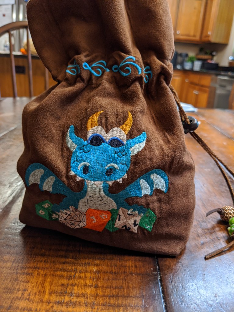 Image of a brown dice bag with a blue embroidered dragon with gold horns happily holding onto its green, white, and orange role playing dice.