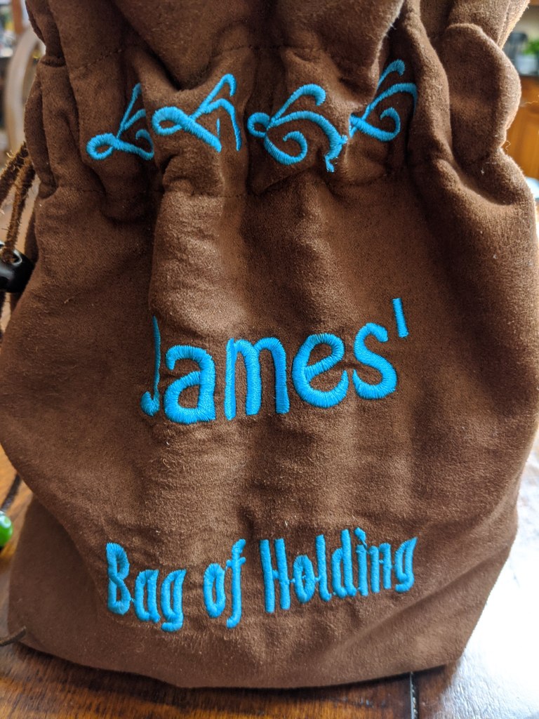 Reverse side of the blue dice bag with the words "James' bag of holding" embroidered on it.