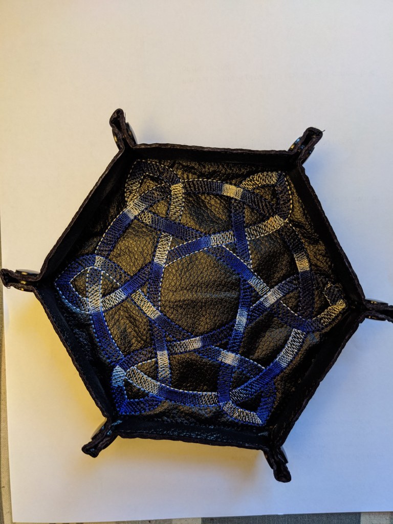 Soft sided dice tray shown in black vinyl with a varigated blue celtic knot embroidered on it.