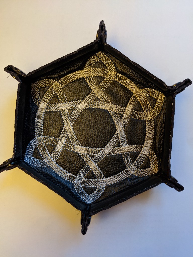 Soft sided dice tray shown in black vinyl with a silver metallic celtic knot embroidered on it.
