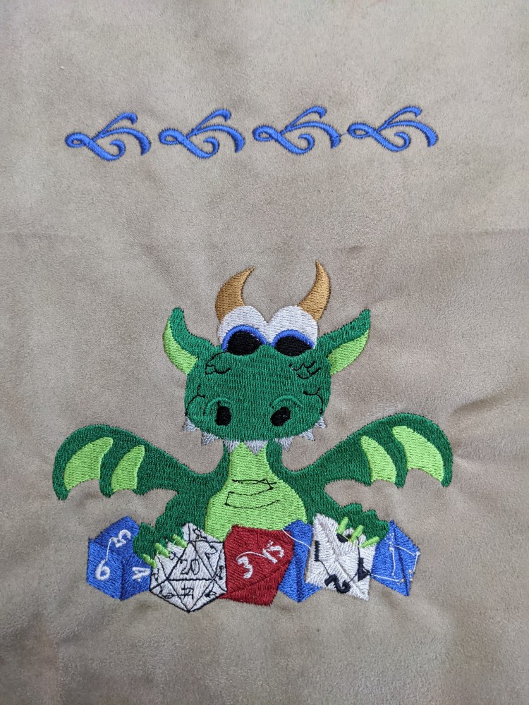 Image is of an embroidered green dragon happily holding onto it's blue, white, and red multi-sided dice.