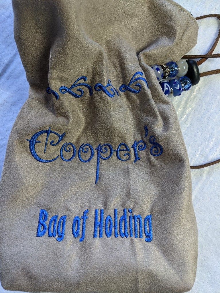 Reverse side of the tan dice bag with the words Cooper's bag of holding embroidered on it.