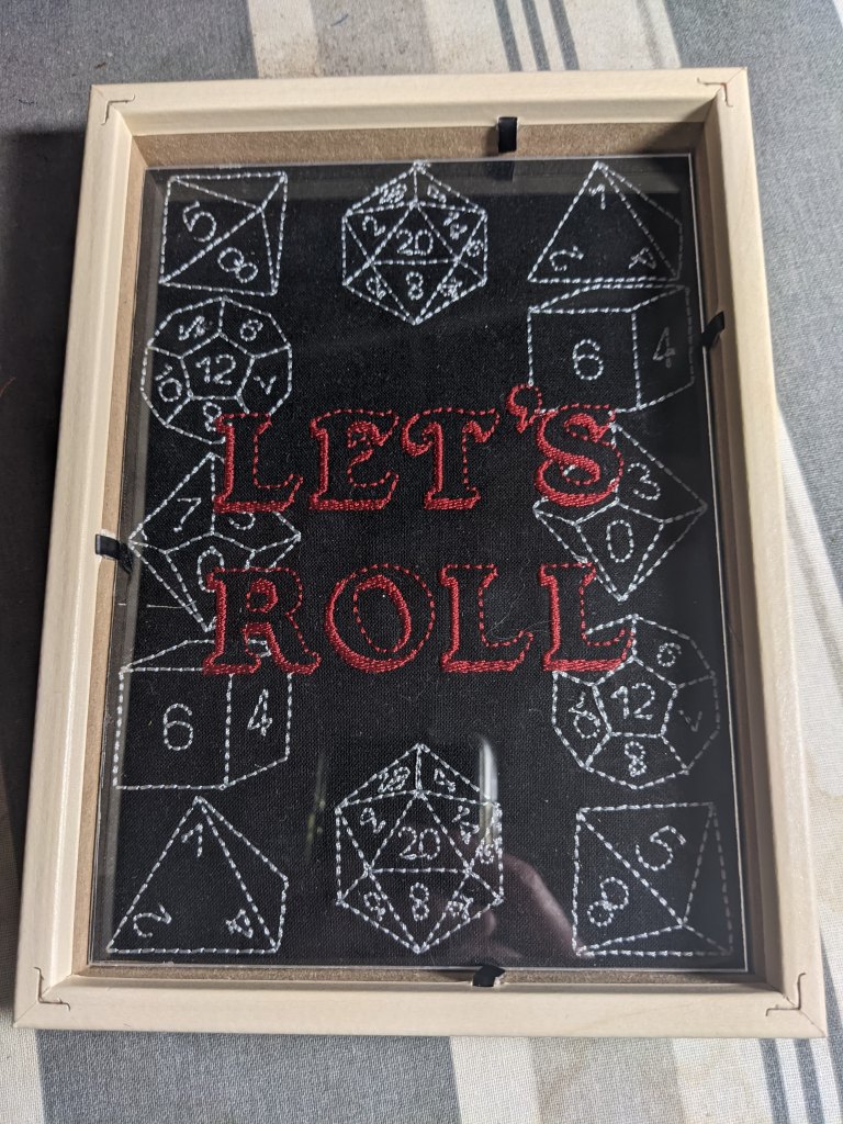 Hard sided dice tray shown in tan frame with black embroidered fabric.  The embroidery says Let's Roll and is surrounded by 4, 6, 12, and 20 sided embroidered dice.